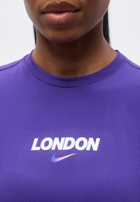 Κοντινό πλάνο ενός ατόμου που φοράει μωβ πουκάμισο με το κείμενο "LONDON" και πολύχρωμο λογότυπο Nike swoosh κάτω από το κείμενο.