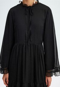 Robe noire à manches longues, avec un col volanté et un détail à nouer à l'avant. Le tissu est lisse avec une jupe évasée et plissée.