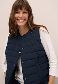Gilet trapuntato blu navy con bottoni a pressione e tasche laterali, indossato sopra una maglietta a maniche lunghe bianca. Texture liscia e design leggero.