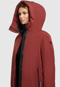 Veste d'extérieur rouge avec une grande capuche, des zips et des accents noirs. Le matériau semble lisse et résistant à l'eau, avec des poches latérales.