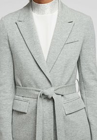 Světlý šedý blazer vyrobený z měkké látky se zavázaným pasem, šálovým límcem a našitými kapsami. Má hladkou texturu a strukturovaný design.