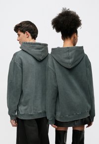 Mikina s kapucňou vo vybledlej tmavozelenej farbe. Má spustené ramená, bočné detaily a rebrované manžety. Mäkká textúra s voľným strihom.