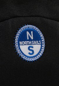 Tessuto nero con una toppa rotonda ricamata con un bordo blu, lettere bianche "NORTH" e "SAILS" e una "N" e una "S" al centro.
