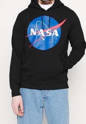 Person wearing black hoodie with large blue NASA logo and light blue jeans, hand in pocket and white shirt visible at neckline.