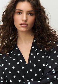 Fekete szövet felsőrész fehér pöttyökkel, V-nyakú kivitelben és hosszú ujjakkal. Sima textúrájú és kényelmes szabású.