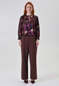 Donna con capelli rossi che indossa una blusa a fantasia viola e marrone, pantaloni larghi marroni e scarpe appuntite borgogna, in piedi su uno sfondo bianco.