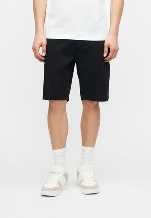 Mann trägt schwarze knielange Shorts, weiße Sportsocken, weiße und beige Turnschuhe sowie ein weißes T-Shirt und steht mit entspannt an den Seiten hängenden Armen.