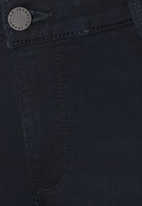 Gros plan sur un jean en denim bleu foncé avec des coutures visibles et un bouton en métal gravé "AG JEANS."