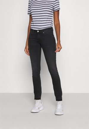 Osoba stojąca, ubrana w czarne jeansy, szaro-białą paskowaną koszulkę z krótkim rękawem oraz białe trampki na jednolitym tle.