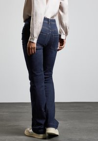 Jean en denim bleu foncé avec un design à jambe droite, dotés de deux poches arrière et d'une taille classique. Portés avec un blouse de couleur claire et des baskets.
