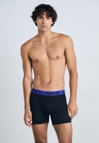 Boxer-briefs noirs avec une large ceinture violette portant le logo "Nike" affiché de manière proéminente à l'avant. Texture lisse et design ajusté.