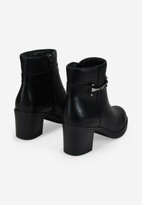 Par de botas de cuero negro hasta el tobillo con tacones de bloque, detalles de hebilla plateada y cremalleras laterales, mostradas desde la parte trasera sobre un fondo blanco.