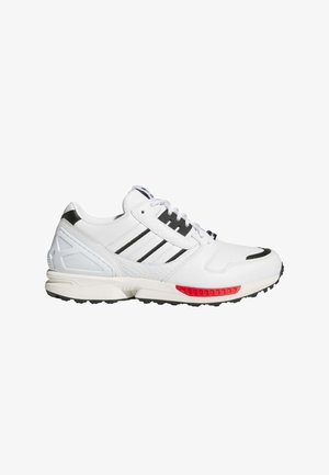 Zapatilla deportiva blanca con rayas negras, acento rojo en la suela, materiales texturizados, diseño con cordones y collar acolchado en el tobillo.