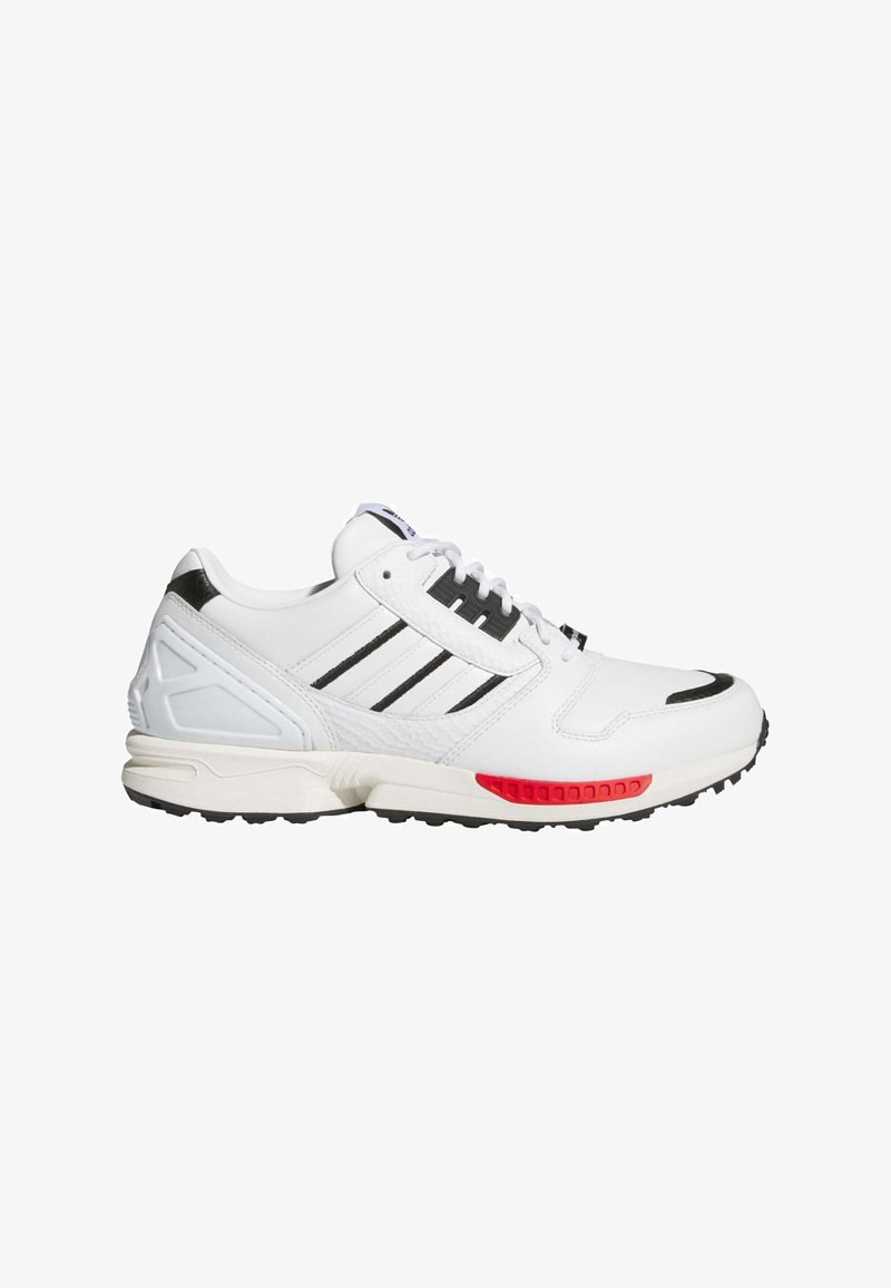 Zapatilla deportiva blanca con rayas negras, acento rojo en la suela, materiales texturizados, diseño con cordones y collar acolchado en el tobillo.