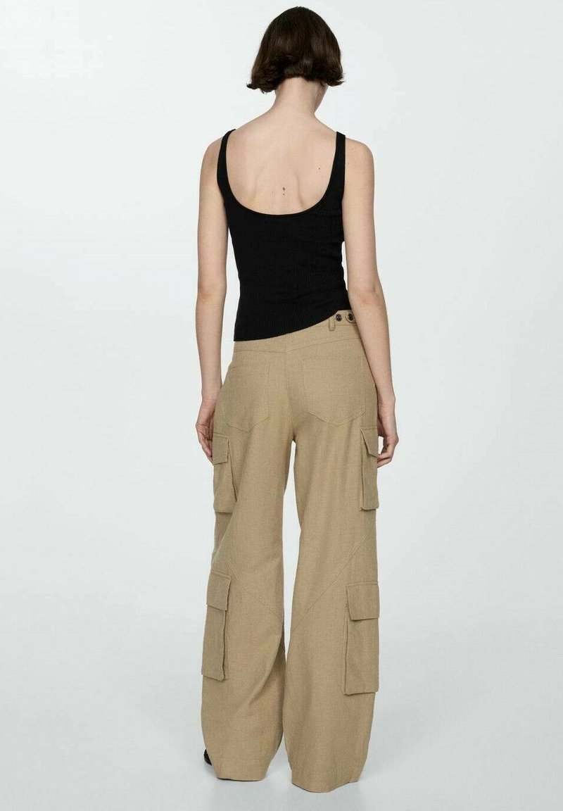 Kobieta z krótkimi brązowymi włosami nosi czarną bezrękawnik i beżowe szerokie spodnie cargo, stojąc tyłem na jednolitym tle.