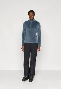 Maglione in pile azzurro chiaro con zip e collo alto, indossato con pantaloni neri e scarponi da sci, che mette in risalto la trama liscia e il design aderente.