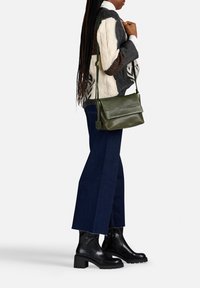 Borsa a tracolla verde in pelle con chiusura a pattina, texture liscia e tracolla regolabile, abbinata a un maglione lavorato a maglia grigio e bianco e jeans scuri.