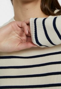 Maglione a coste color crema con strisce blu navy. Presenta polsini aderenti e colletto rotondo. La texture è morbida ed elastica.