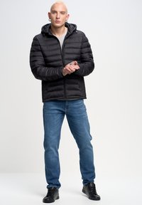 Sort pufferjakke med vandret syninger, lynlåslukning og hætte, parret med lyseblå jeans og sorte sneakers.