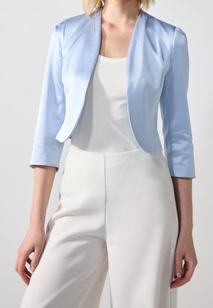 Femme porte un blazer court bleu clair avec des manches trois-quarts sur un haut blanc et un pantalon blanc taille haute sur un fond gris.