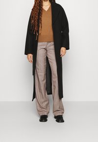 Black long coat over a brown knit v-neck sweater, paired with wide plaid trousers in beige and brown. Black shoes complete the outfit.