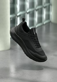 Μαύρο αθλητικό παπούτσι Calvin Klein με υφή στα πλευρά και κορδόνια, αιωρούμενο πάνω από γυαλισμένο δάπεδο από σκυρόδεμα κοντά σε τζάμια κυψελωτού τύπου.