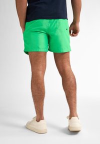 Șorturi de baie verzi cu o textură netedă și un buzunar la spate, asortate cu adidași de culoare deschisă. Design minimalist care pune accent pe funcționalitate.