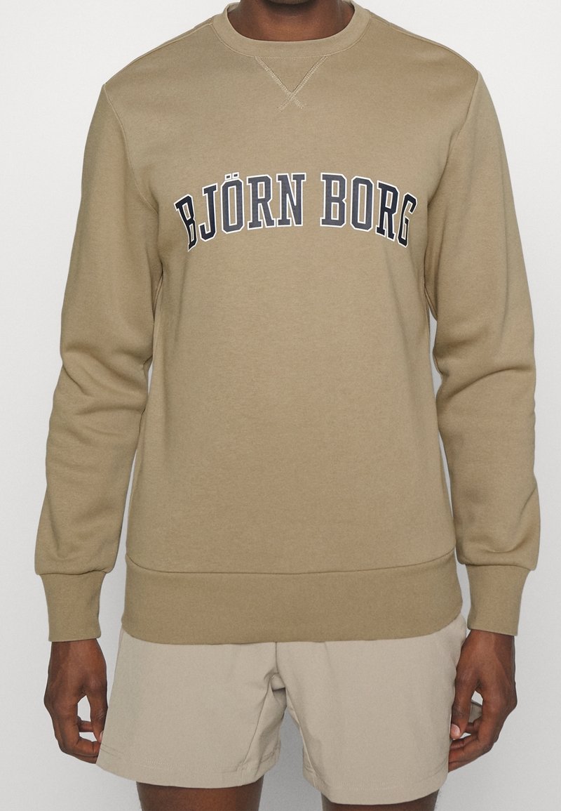 Sweat-shirt beige avec des poignets et un ourlet côtelés, arborant le texte "BJÖRN BORG" imprimé en bleu marine sur la poitrine. Matière en mélange de coton doux.