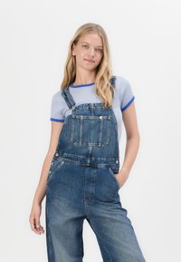 Jeans-Overalls in Mittelblau mit einer Vordertasche, verstellbaren Trägern und silberfarbenen Beschlägen, kombiniert mit einem blau-weißen gestreiften Hemd.
