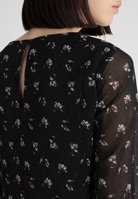 Svart blus med blommönster som har en nyckelhålssnitt bak, genomskinliga ärmar och en strukturerad tyg med små vita och röda blommor.