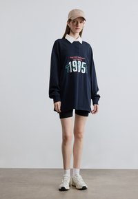 Λευκός φαρδύς φούτερ του Ναυτικού με λευκό γιακά και τυπωμένο λογότυπο, σε συνδυασμό με μαύρο σορτς και λευκά αθλητικά παπούτσια, ανοιχτό μπεζ καπέλο μπέιζμπολ.