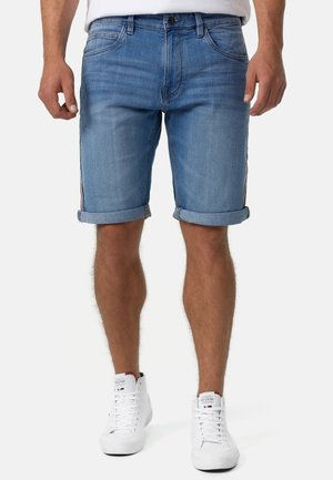 Mand iført opsmøgede blå denimshorts og hvide høje sneakers stående mod en ensfarvet baggrund.