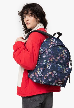 Mochila con patrón floral en azul marino, con correas ajustables y un bolsillo frontal, hecha de tela duradera. El modelo lleva una chaqueta roja y jeans grises.