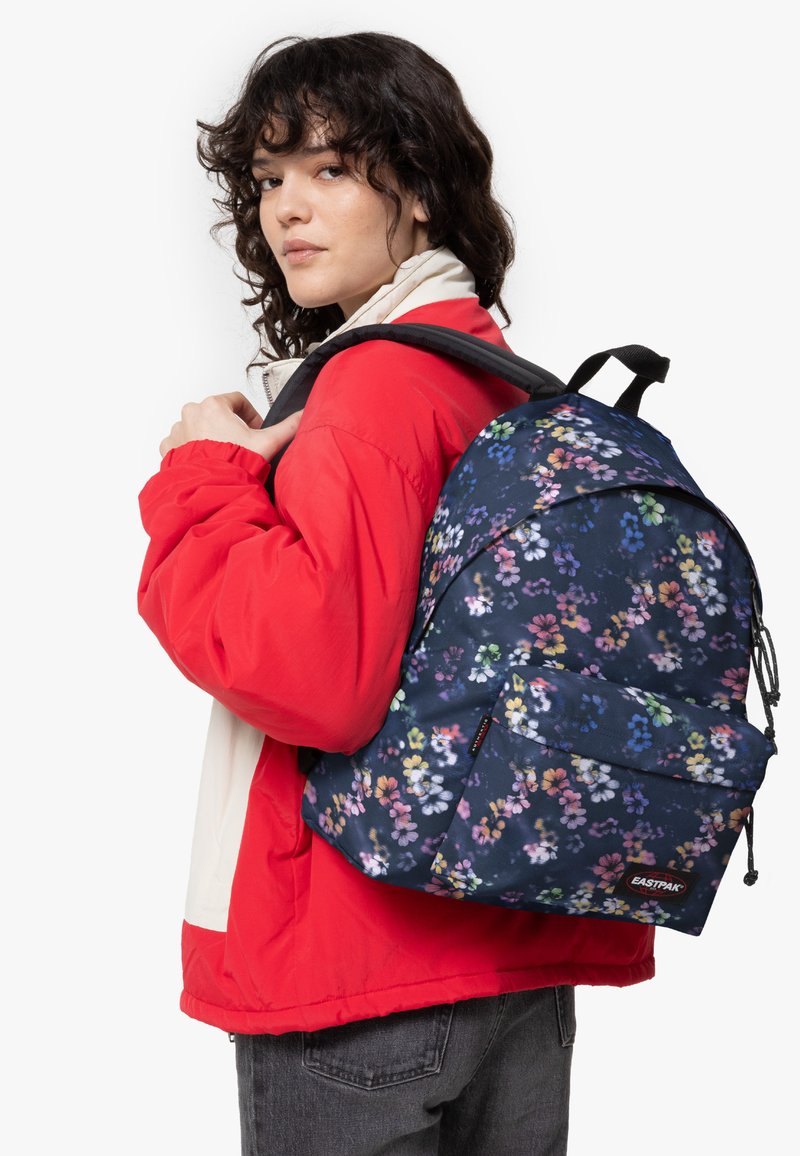 Zaino blu con motivo floreale, dotato di spalline regolabili e tasca anteriore, realizzato in tessuto resistente. Il modello indossa una giacca rossa e jeans grigi.