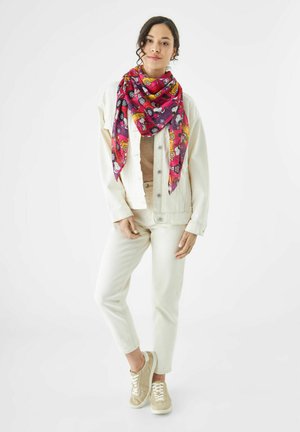 Écharpe en soie colorée avec un motif rose et violet, comportant des personnages de dessins animés. Le mannequin porte une veste en denim crème, un haut beige et un pantalon clair.