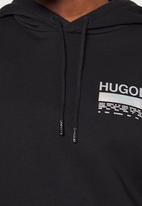Zwarte hoodie van zachte stof, met trekkoorden en een reflecterend logo met tekst aan de voorkant, met de nadruk op modern design en comfort.
