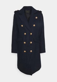 Cappotto doppiopetto blu navy con bottoni decorativi dorati e mostrine sulle spalle, lunghezza fino al ginocchio con tasche frontali a pattina.