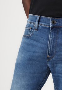 Blå denimjeans med en slät textur. Har en knapp- och dragkedjestängning, bältesöglor och två framfickor.