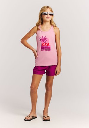 Roze katoenen tanktop met een grafische opdruk "Summer Daze", gecombineerd met paarse shorts. Zwarte slippers en een zonnebril maken de outfit compleet.