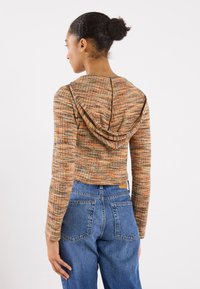 Top corto con cappuccio in tessuto testurizzato multicolore con tonalità di arancione e marrone. Indossato con jeans di denim blu a vita alta.