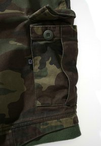 Tela con patrón de camuflaje que presenta tonos verdes, marrones y negros, con un botón negro y un detalle de bolsillo lateral. Material suave y texturizado.