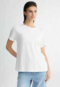 Biała bawełniana koszulka z okrągłym dekoltem, krótkimi rękawami i ozdobną kieszenią z koralikami. Casualowy krój, minimalistyczny design.