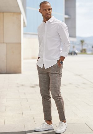 Weißes Langarmhemd, gemusterte beige Hose und weiße Sneakers. Modell steht im Freien mit Händen in den Taschen und präsentiert legere Kleidung.