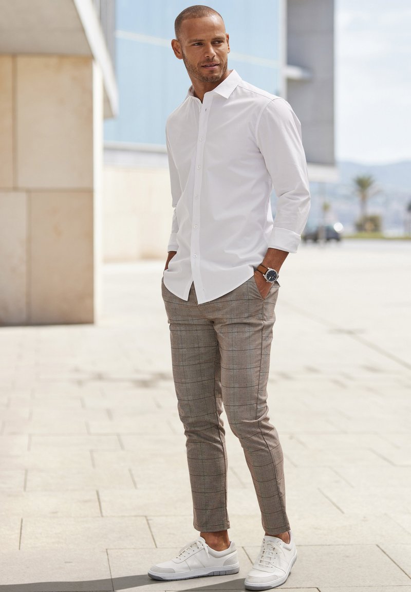 White long-sleeve shirt, patterned beige trousers, and white sneakers. Model standing outdoors with hands in pockets, displaying casual attire.