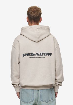 Personne portant un sweat à capuche beige avec le texte « PEGADOR Certified Authentic Streetwear » au dos, associé à un jean bleu clair.
