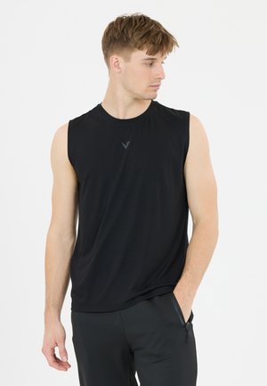 Junger Mann trägt schwarzes ärmelloses Shirt und schwarze Hose, eine Hand in der Tasche, schaut nach links.