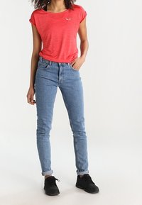 Červené tričko s krátkým rukávem a malým logem na prsou, v kombinaci se světlými, přiléhavými modrými džínami a černými teniskami. Jednoduchý, ležérní outfit.