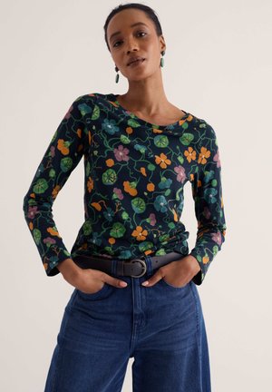 Frau trägt ein dunkel geblümtes Langarmshirt, blaue Jeans, einen schwarzen Gürtel und grüne Hängeohrringe und steht mit den Händen in den Taschen.