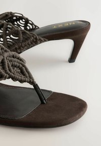 Brun ruskindsandal med høje hæle, fletede remme og en tånenskeform på hvid baggrund.