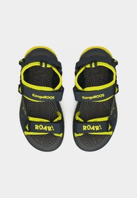 Παιδικά σανδάλια Kangaroos σε μαύρο και κίτρινο χρώμα με λουριά βέλκρο, ανάγλυφο μοτίβο στη σόλα και το κείμενο "ROAR!" στα μπροστινά λουριά.